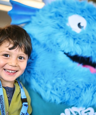 Person standing next to a large blue furry mascot with white eyes and a big smile in the Matrosklubben.