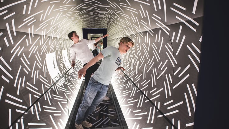 Two people walking through an illuminated tunnel with white line patterns.