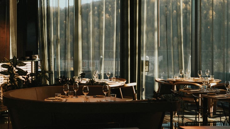 Elegant restaurant interior with tables set for dining and forest view through large windows.