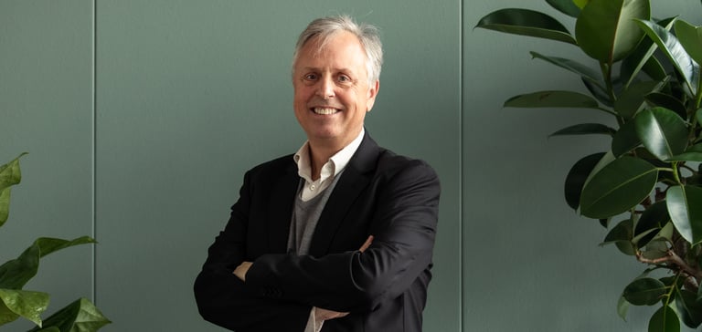 Person standing indoors with arms crossed, wearing a dark suit jacket and surrounded by green plants.