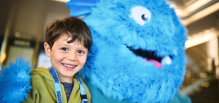 Person standing next to a large blue furry mascot with white eyes and a big smile in the Matrosklubben.